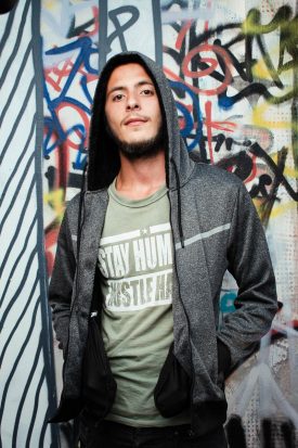 Portrait of refugee Mohammad with his hands in the pockets of his grey hoodie standing against a graffiti wall