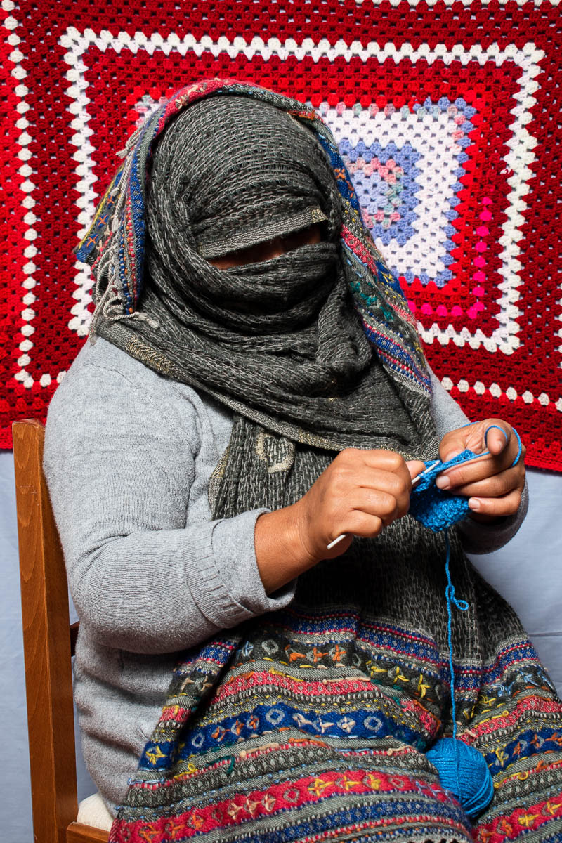 Portrait of refugee Arezo knitting while wearing a woollen scarf that covers all her face and head