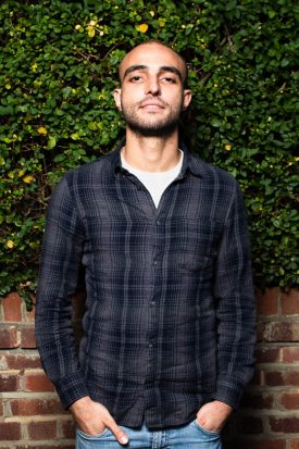 Portrait of refugee Zuheir wih his hands in his pocket against a hedge