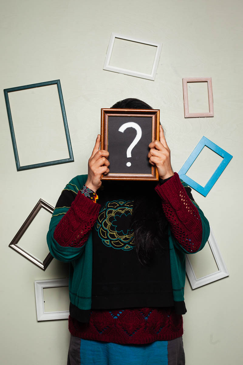 Portrait of refugee Axiom surrounded by blank wall frames holding a frame with a question mark to hide their face