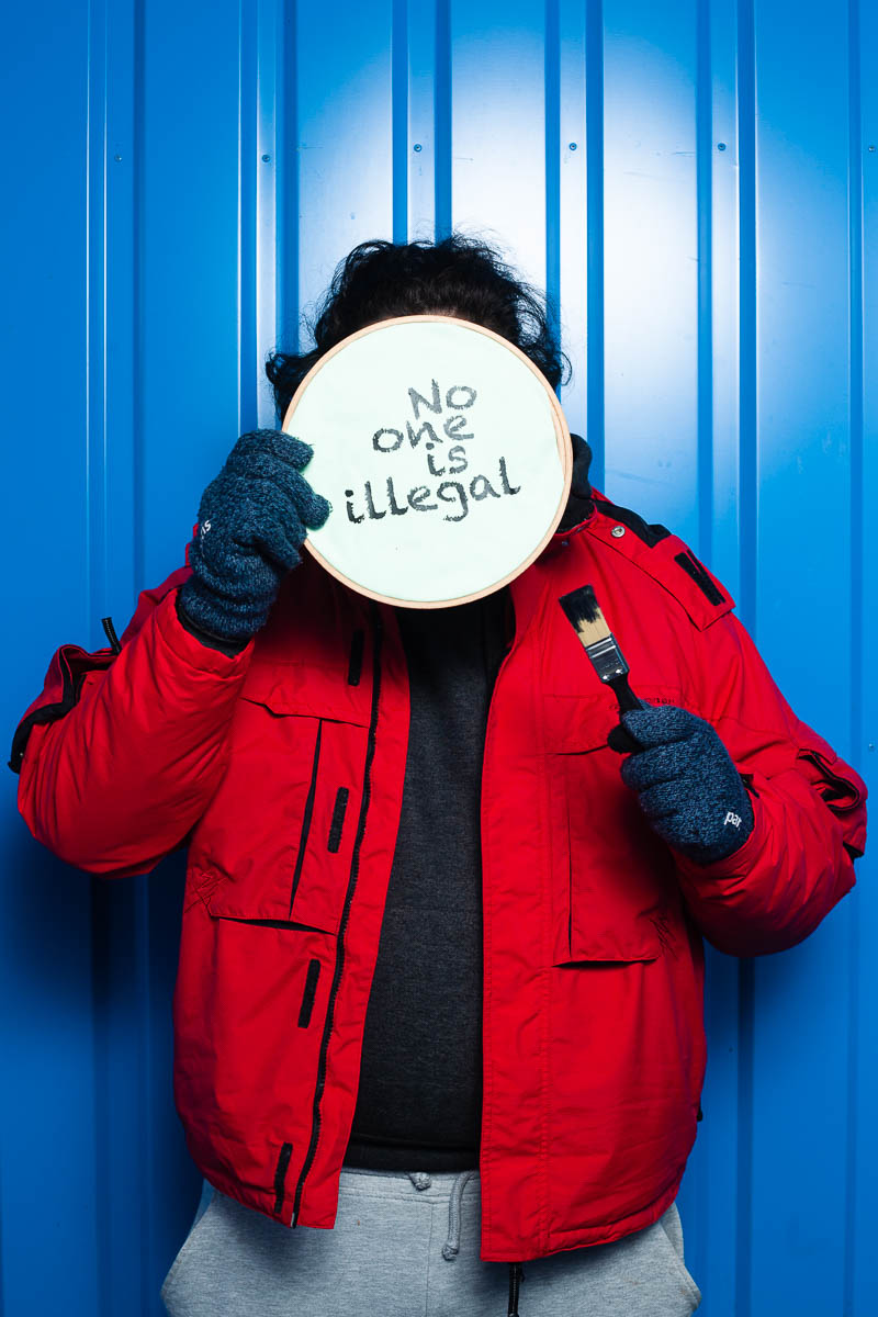 Portrait of refugee Morrissey hiding face with a canvas saying "no one is illegal"