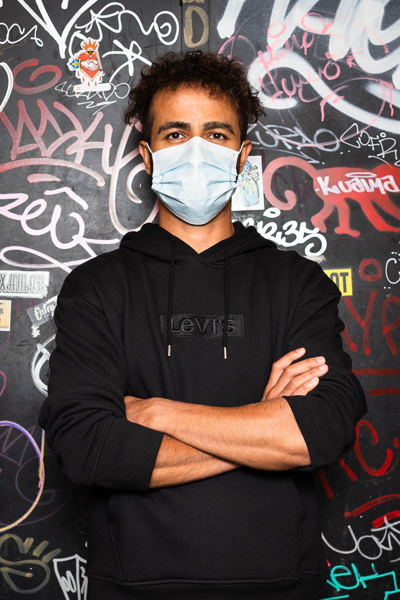 Portrait of refugee Serken Kilic wearing a mask and folding his hands against a graffiti background