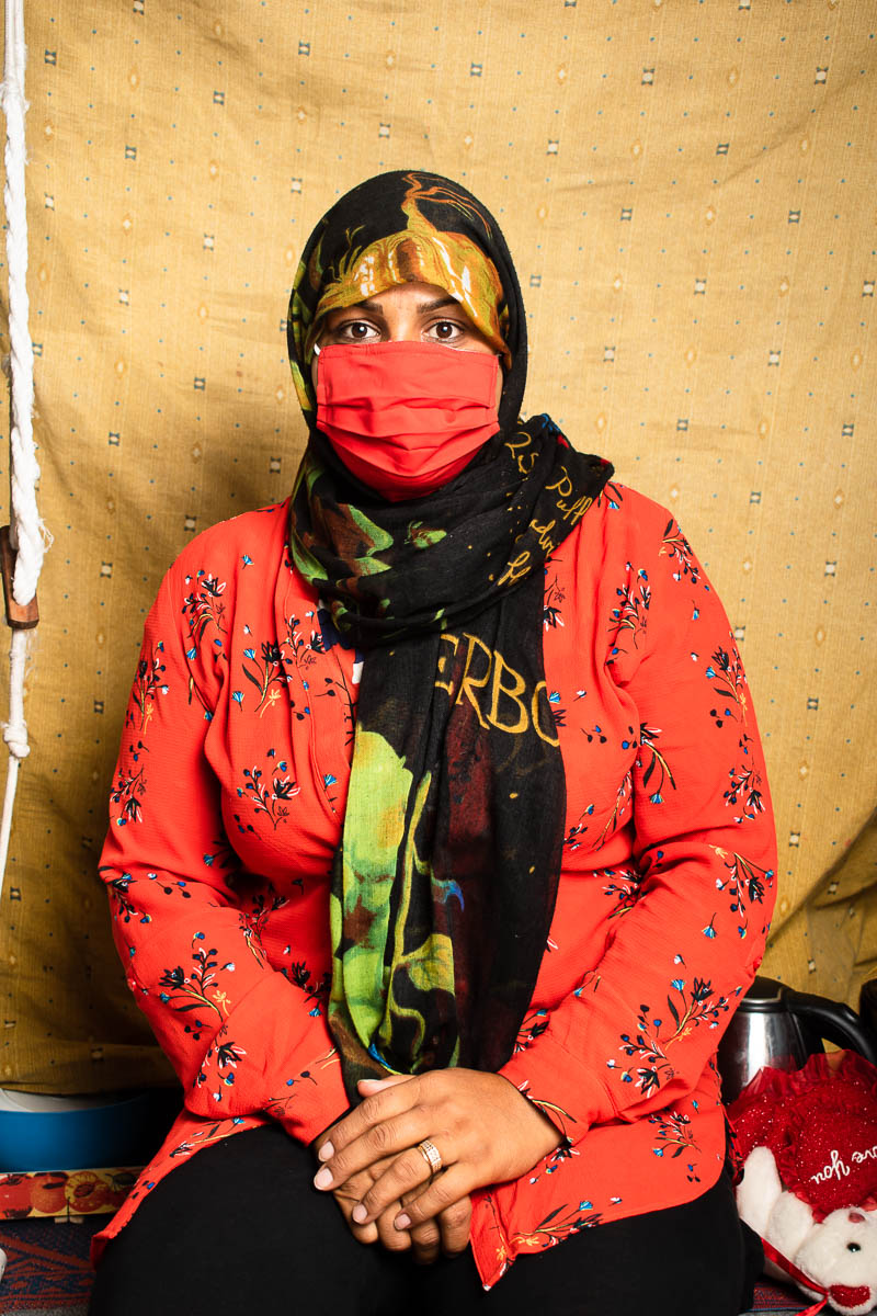 Portrait of refugee Samira in a hijab and red mask sitting down.