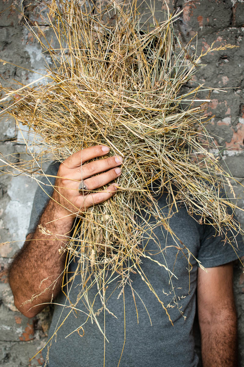 Portrait of refugee Ashkan hiding his face with a bunch of dried grass