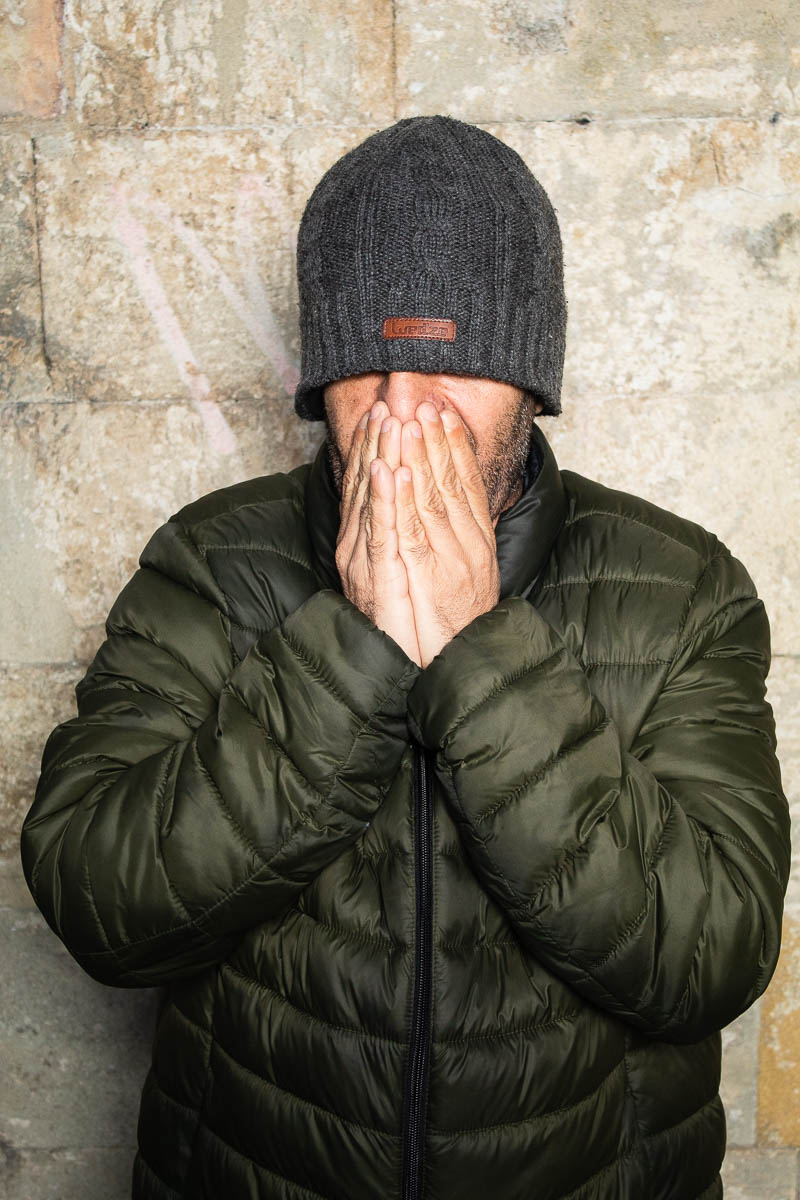 Portrait of refugee Zhiva wearing a woollen cap coving their eyes and hands covering the face