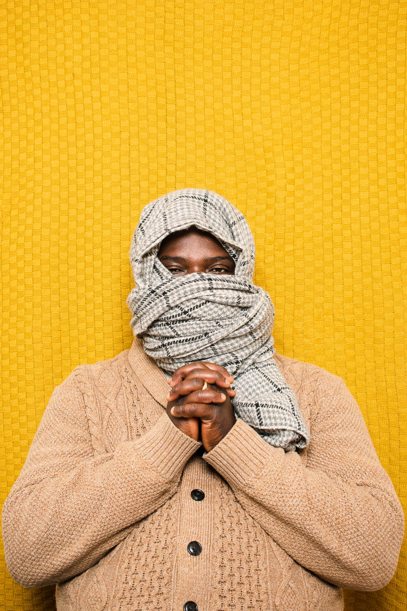 Portrait of refugee Kodjo holding hands interlocked together with a scarf covering their head and half the face
