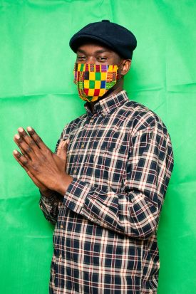 Portrait of refugee Tchok Le maitre holding his folded hands wearing a colorful embroidered face mask and a green beret