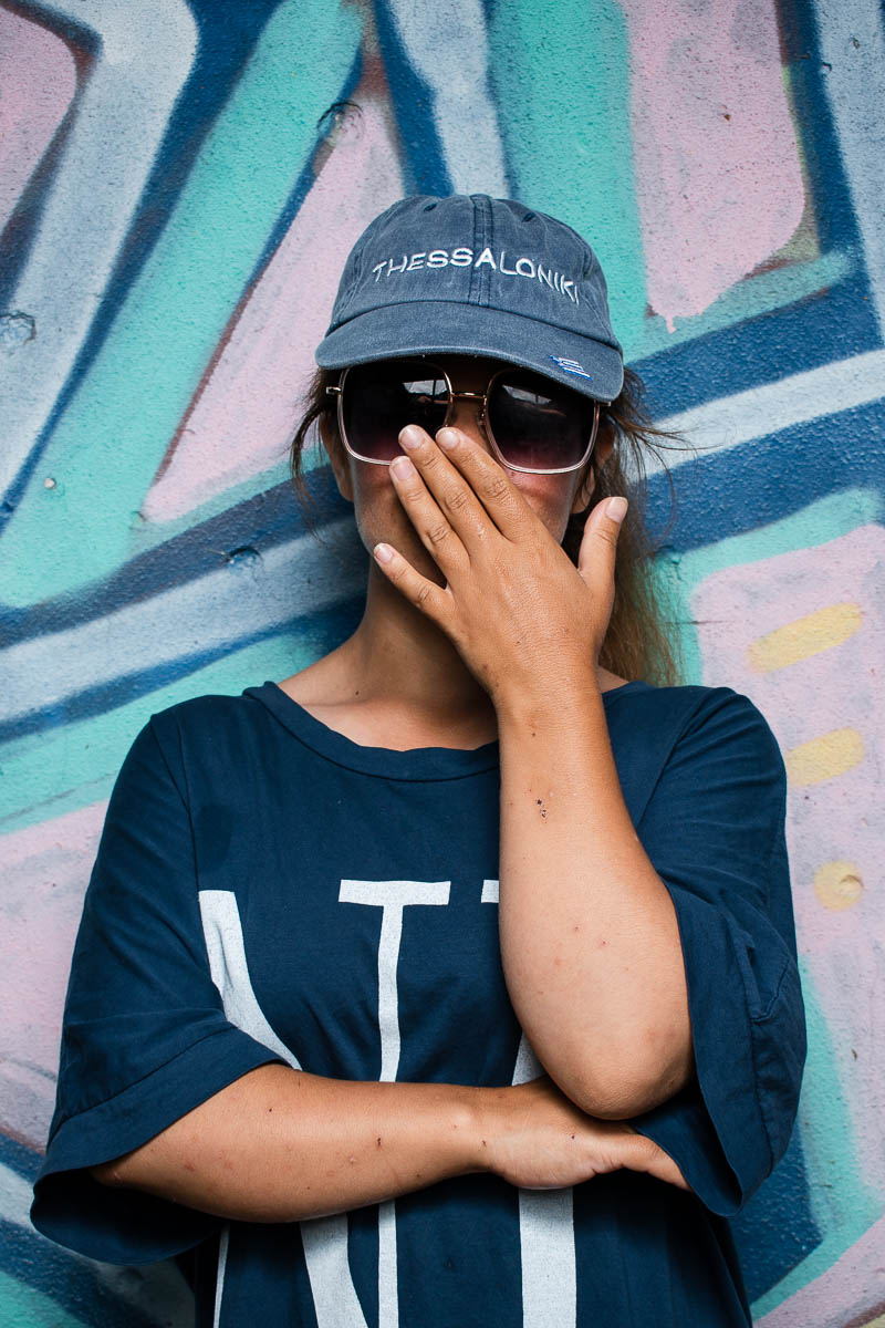 Portrait of refugee Mahsa wearing shades and a cap, hiding her face and standing against a graffiti wall