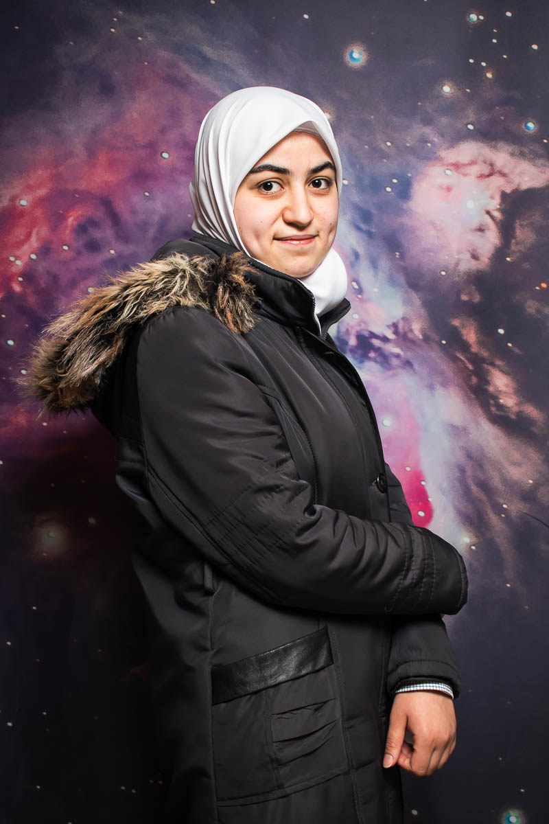Portrait of refugee Riham standing sideways wearing a parka and a hijab