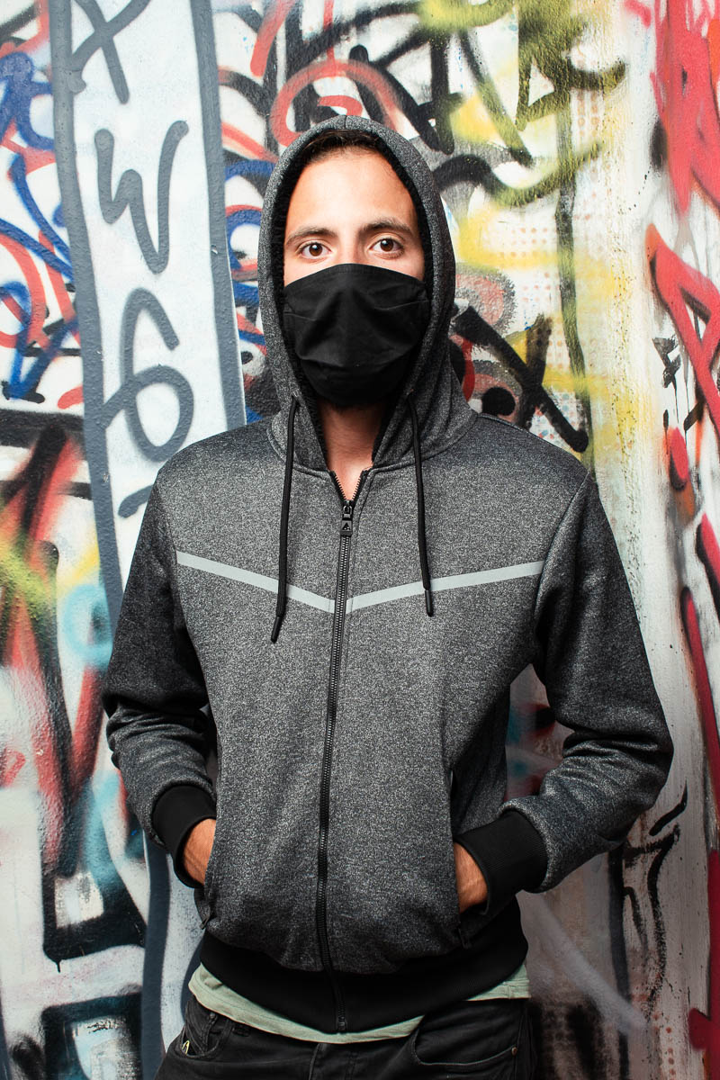 Portrait of refugee Mazen wearing a mask and a hoodie with his hands in the front pockets