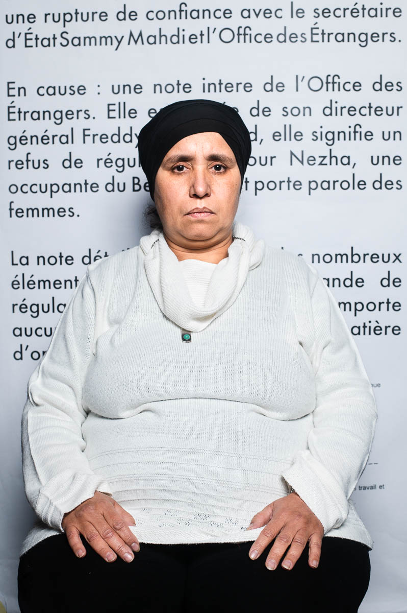 Portrait of refugee Nezha wearing a black head cap against a wall with french writing on it