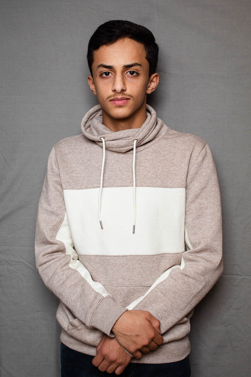 Portrait of refugee Mesbar wearing a hoodie with his hands folded in front of him.