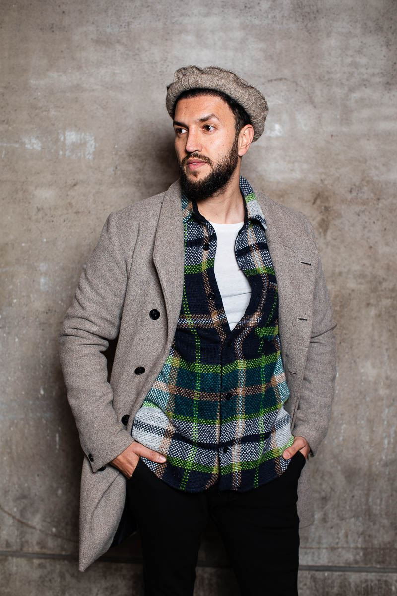 Portrait of refugee Sameer wearing a beret and a plaid vest with his hands in the pockets of his pants looking sideways