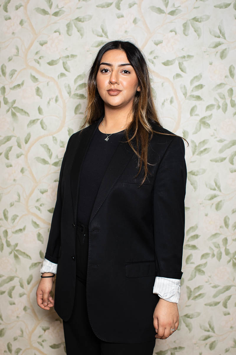 Portrait of refugee Maryam in a suit standing against a floral wallpaper