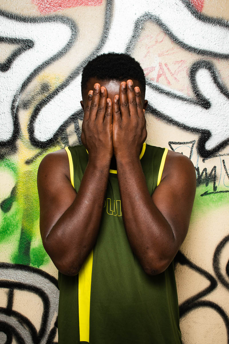 Portrait of refugee Abdulai covering his face with both hands, standing against a graffiti wall