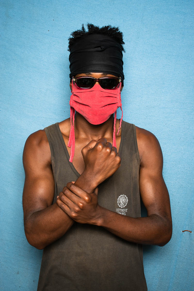 Portrait of refugee Daniel wearing a red mask, shades and a bandana holding a fist and the other hand holding it