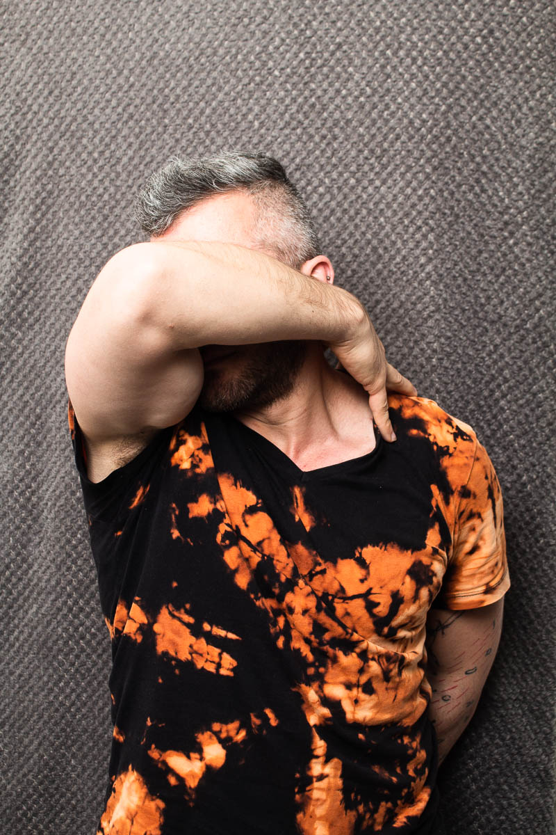 Portrait of refugee Rassaya wearing a dyed shirt with his arm folded across his face to cover it