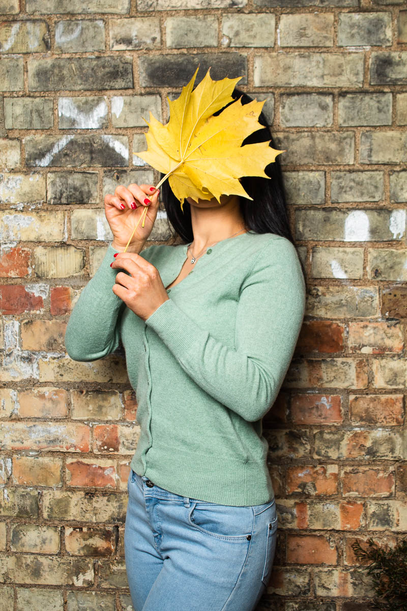 Portrait of refugee Ritta standing against a brick wall, using a large fall leaf to cover her face