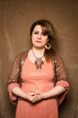 Portrait of refugee Midya wearing an orange dress with gold sequined sleeves holding her hands in front of her