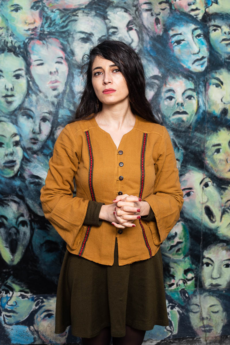 Portrait of refugee Hazel standing against a painted wall portrait of faces
