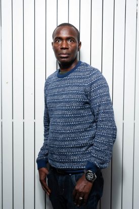 Portrait of refugee Almamy wearing a blue sweater with his thumbs in his jean pocket