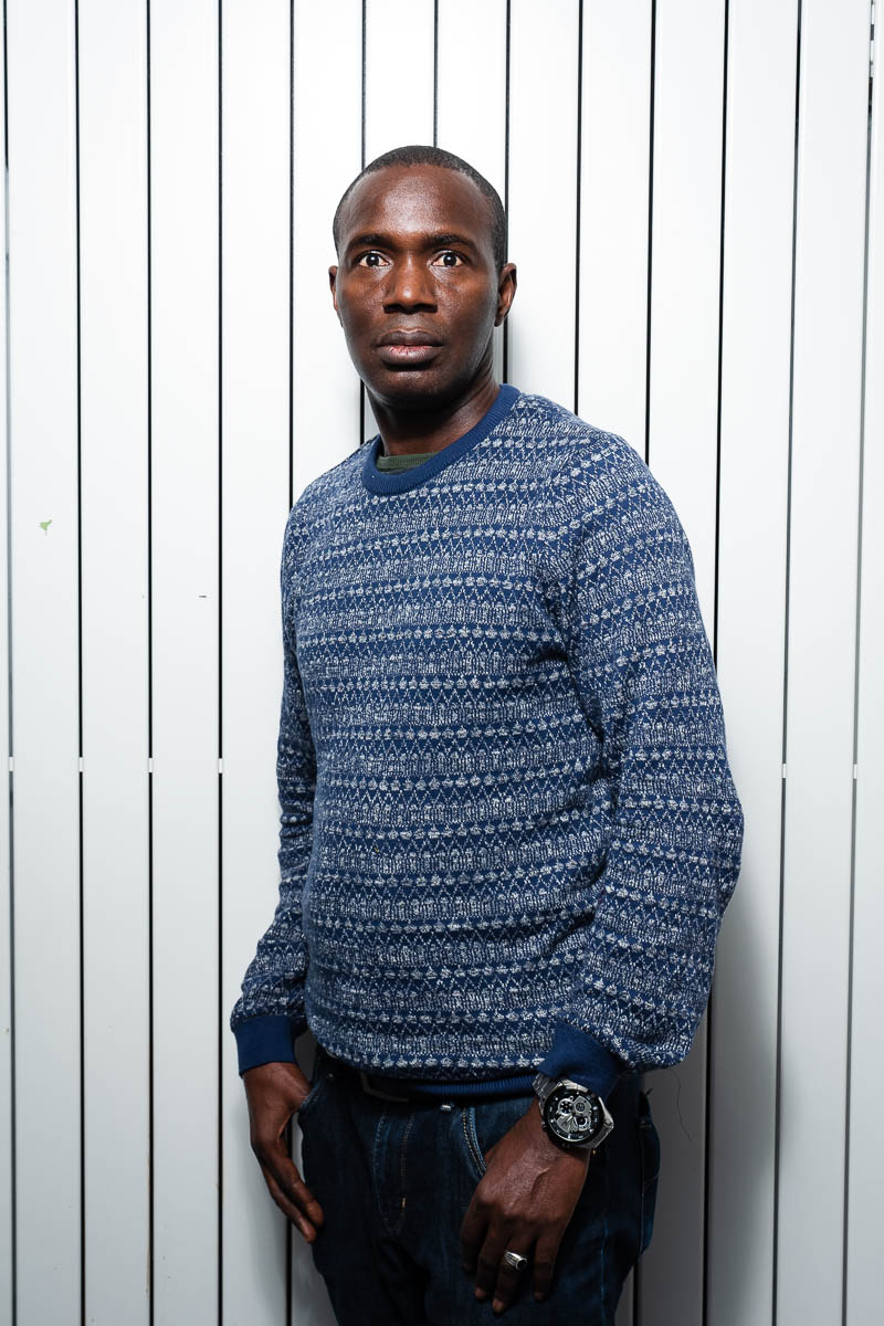 Portrait of refugee Almamy wearing a blue sweater with his thumbs in his jean pocket