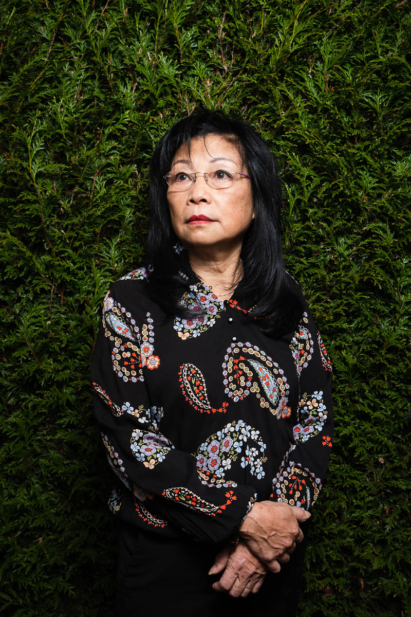 Portrait of refugee Hoang with her hands folded in the front against a green hedge background