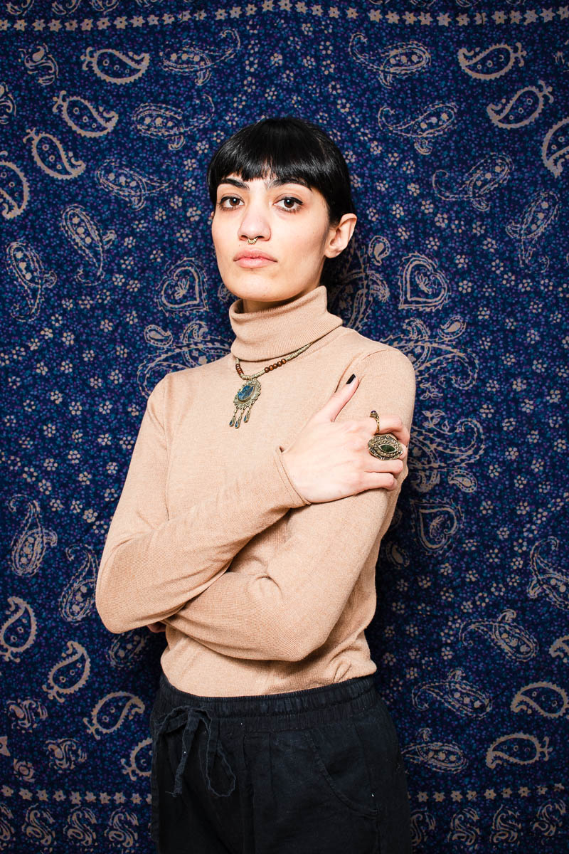 Portrait of refugee Taraneh with her arms wrapped around herself against a blue background with designs