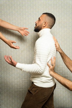 Portrait of refugee Greece turned sideways to the left with his hands raised while two hands support his back and another pair of hands reach out to him towards the front