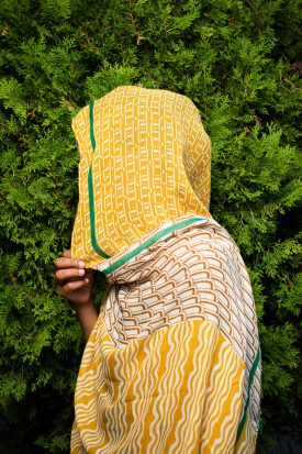 Portrait of refugee Jasmin turned sideways to the left, fully covered with a shawl only her hand holding it against a plant background.