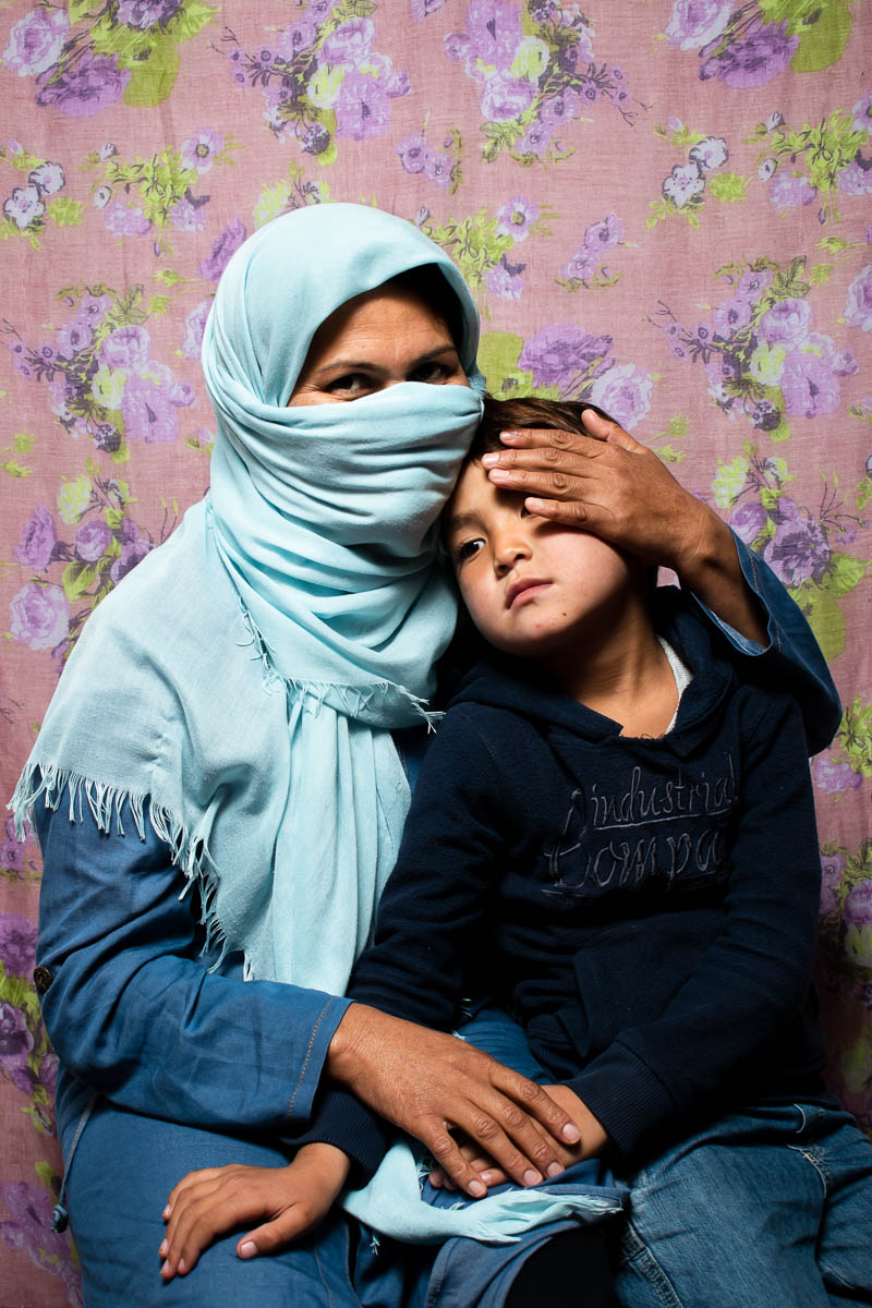 Portrait of refugee Zahra covering her full face and head with a light blue scarf holding a child on her lap