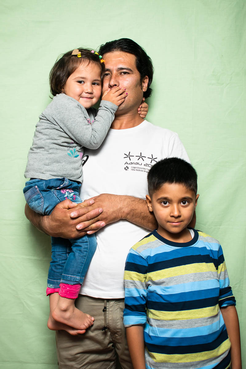 Portrait of refugee Noor holding a little girl whose hands are covering his mouth and a little boy standing to the right