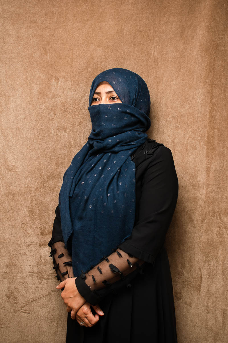 Portrait of refugee Sahar with a blue hijab covering her head and lower half of face, standing with her hands folded to her front
