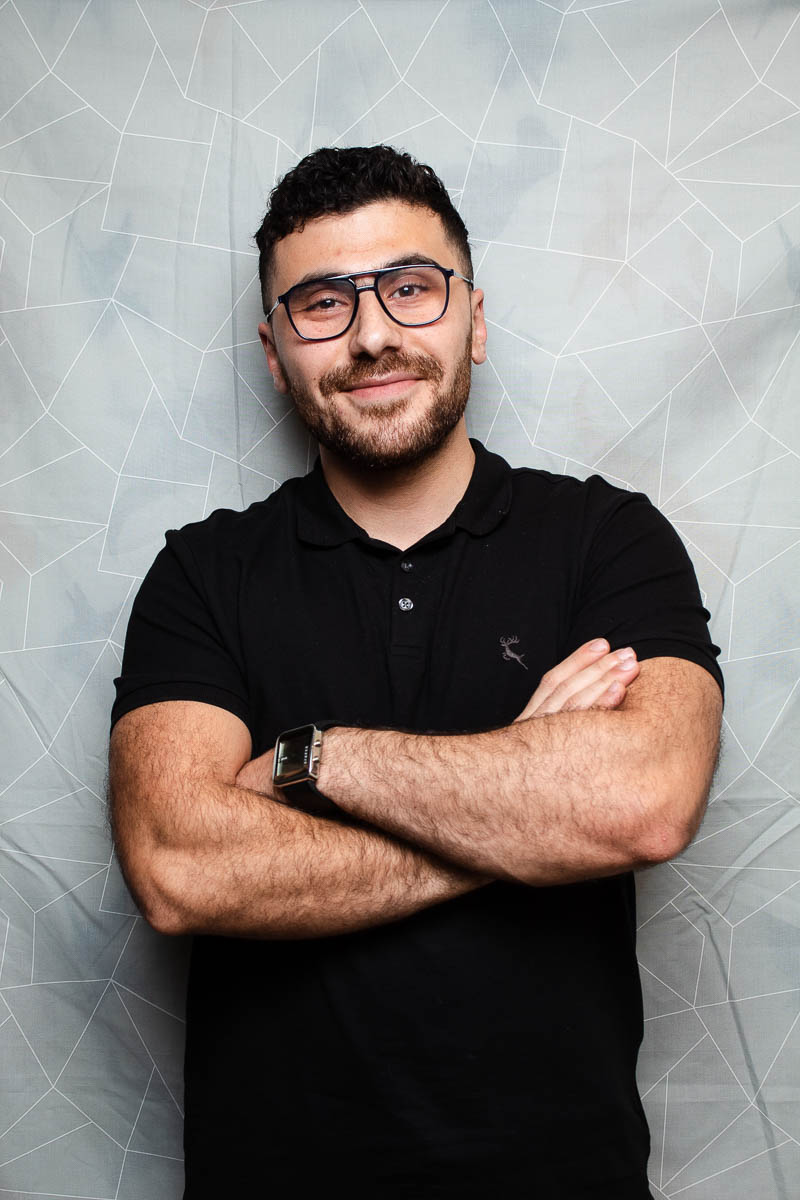 Portrait of refugee Amro wearing a black polo shirt and glasses smiling with his arms crossed across his chest