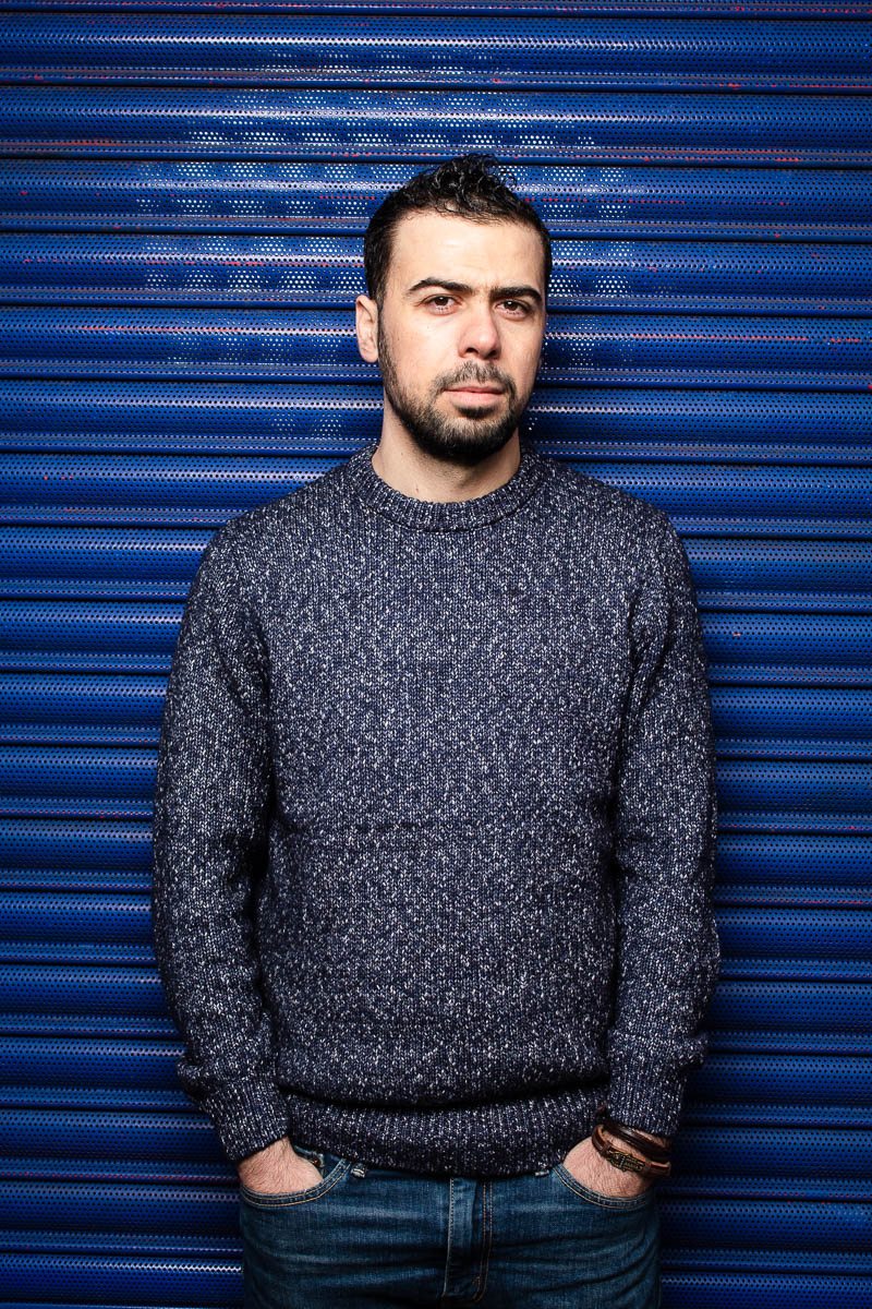 Portrait of refugee Abdullah wearing a sweater with his hands in the pocket of his jeans standing against a blue shutter background