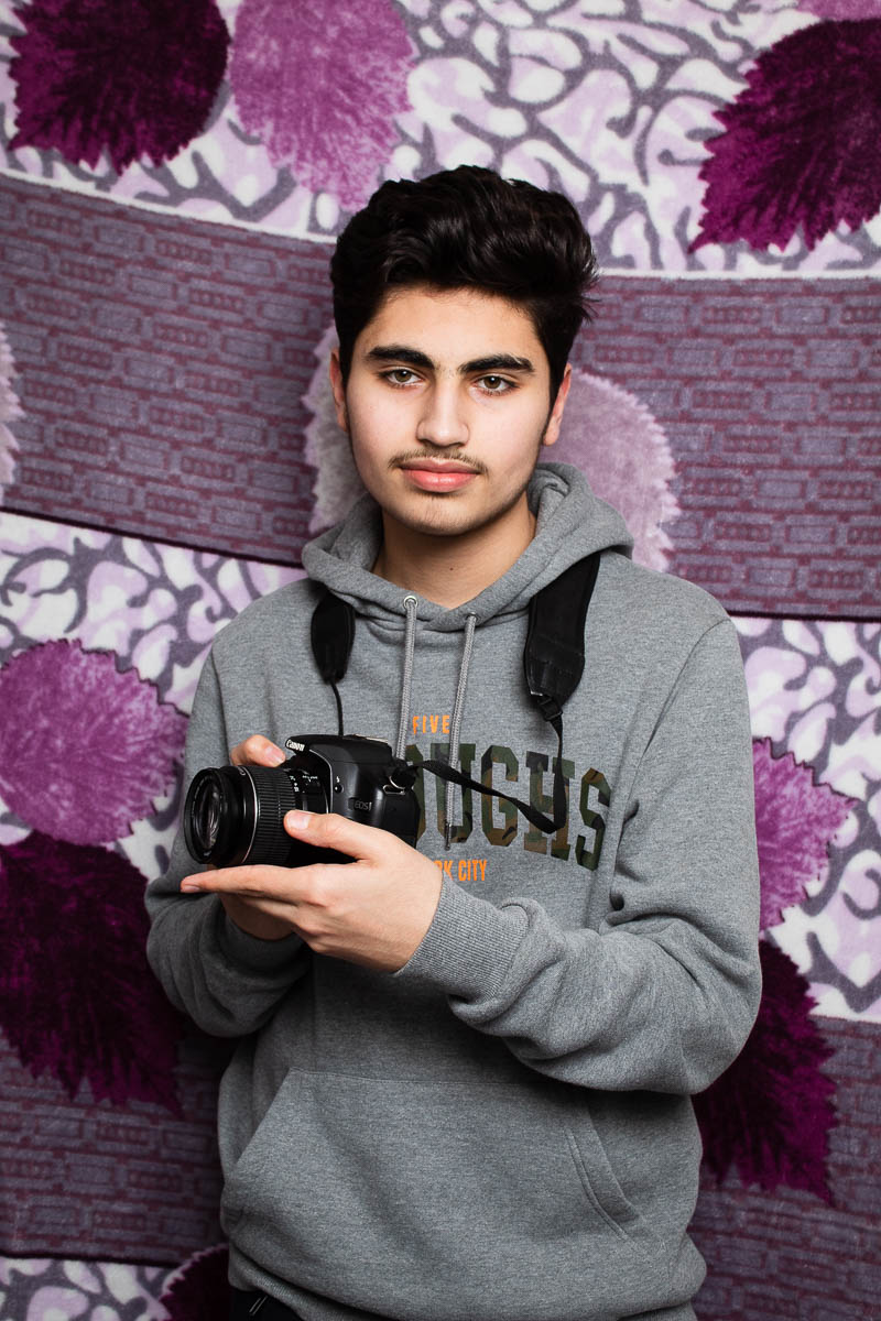 Portrait of refugee Husam wearing a hoodie holding a camera in his hands