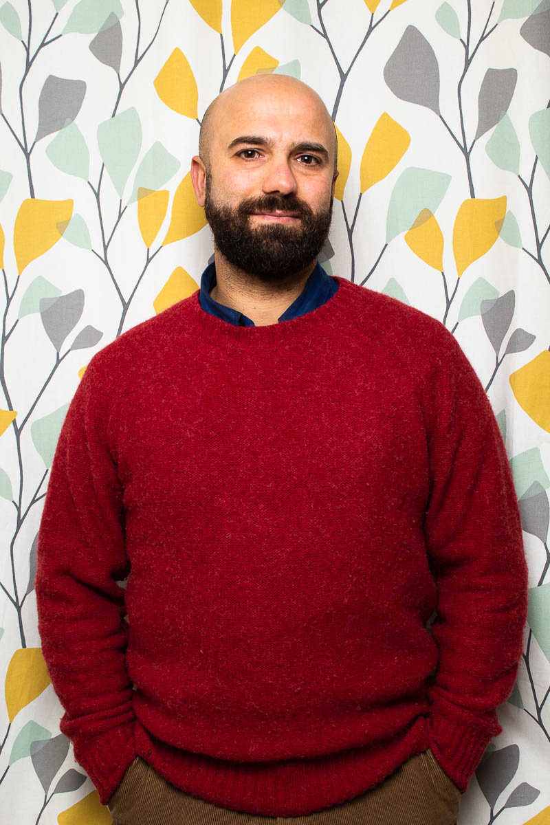 Portrait of refugee Basel with his hands in his pant pockets wearing a red sweater standing against a wall with a colorful floral wallpaper