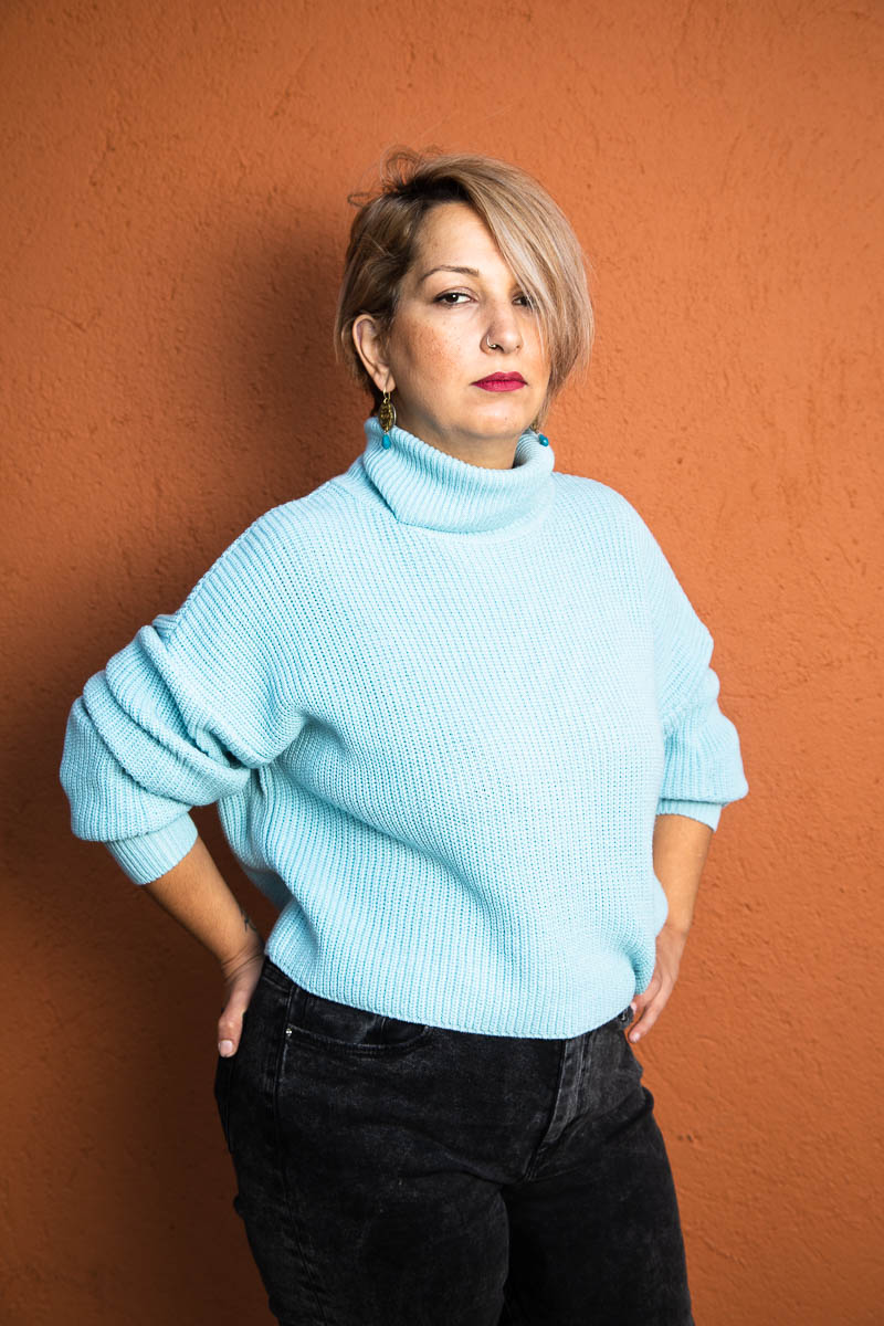 Portrait of refugee Shadi wearing a blue turtleneck sweater standing with her hands in her back pockets