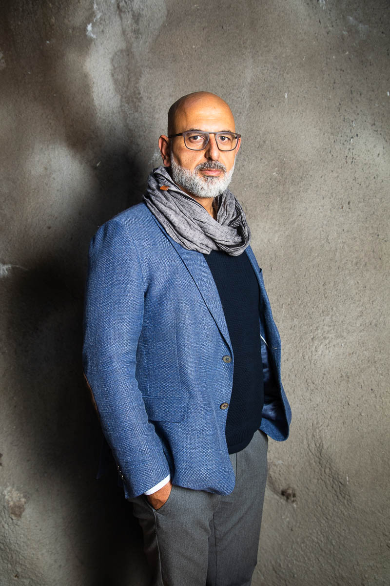 Portrait of refugee Karim standing sideways with his hands in his pockets wearing a grey scarf around his neck and a blue blazer