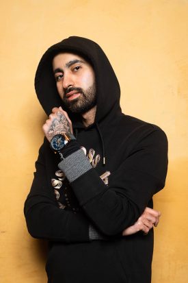 Portrait of refugee Arya wearing a hoodie and holding his left tattooed hand to his chin