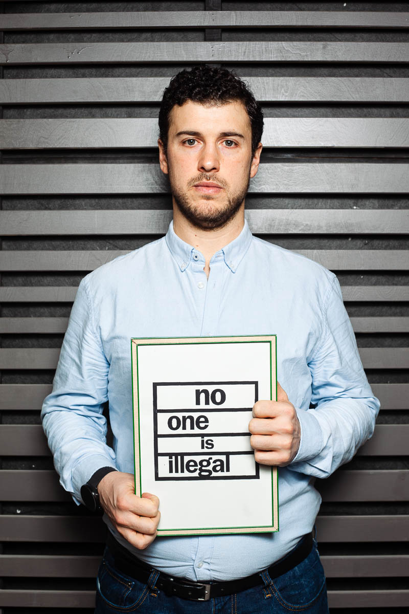 Portrait of refugee Diego standing against a metal screen holding a frame that reads "no one is illegal"