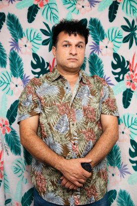 Portrait of refugee Nabaz with his hands on top of the other wearing a shirt with tropical leaves and feathers standing against a wall matching his shirt