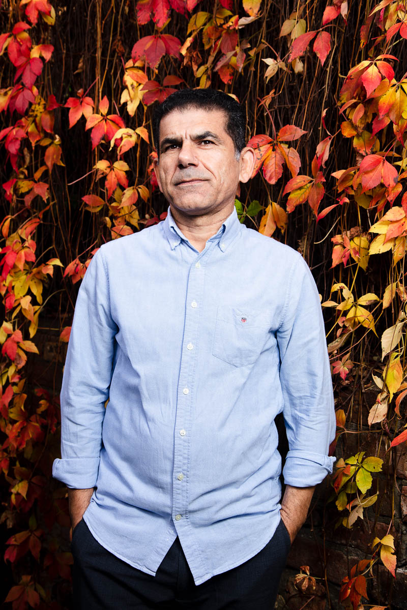 Portrait of refugee Ali with his hands in his pockets standing against a fall leaves background