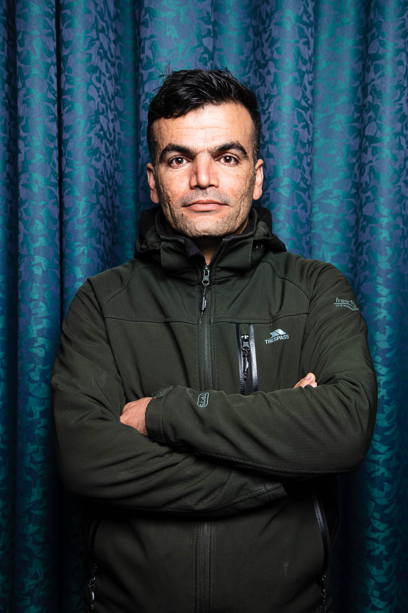 Portrait of refugee Karwan with his arms crossed standing against a blue curtain