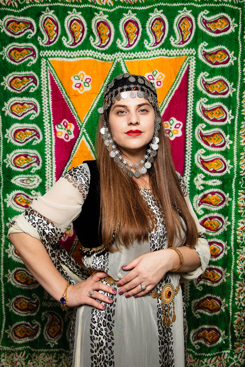Portrait of refugee Asia wearing a kurdish headpiece with her hand oh her hip standing against an embroidered patterned background