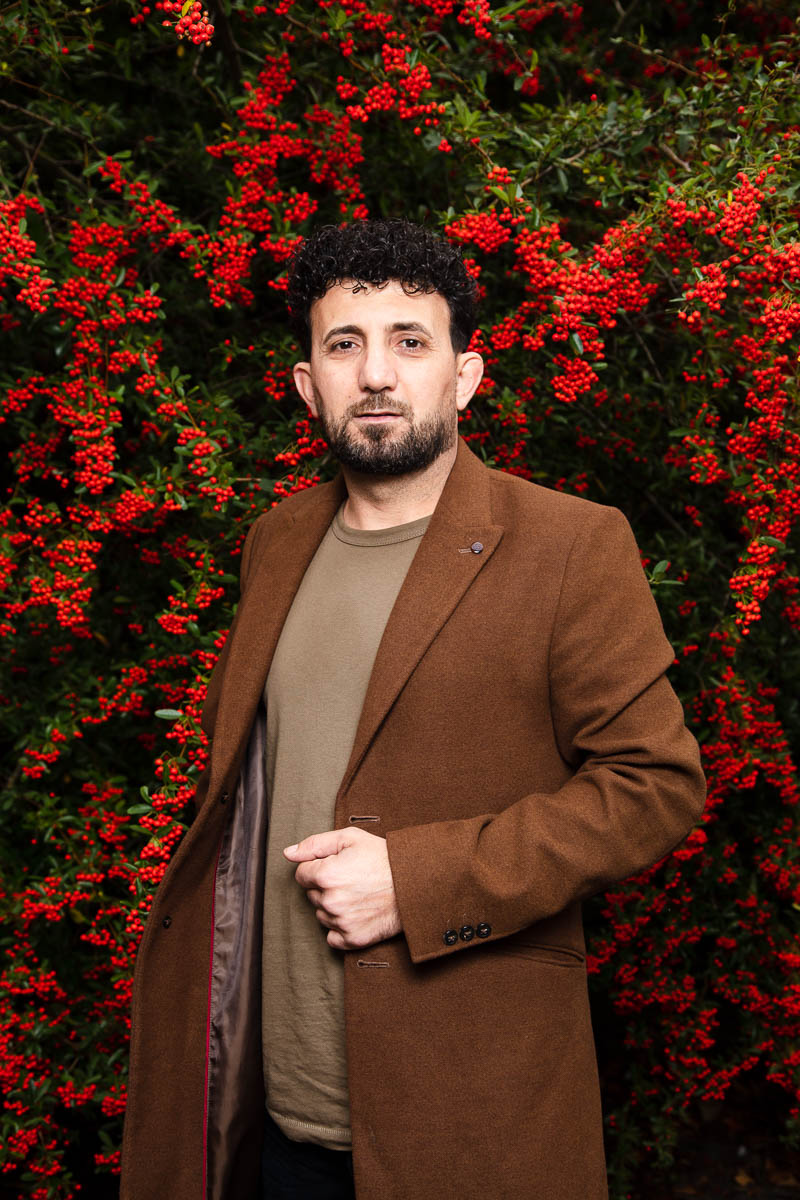 Portrait of refugee Tala holding the lapels of his brown jacket standing against a background with red flowers