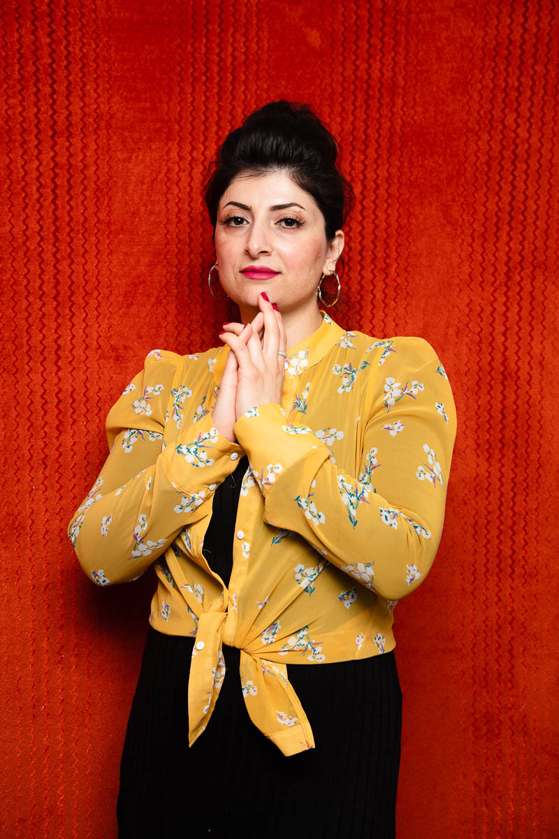 Portrait of refugee Shna wearing a yellow floral cardigan with her hands folded and resting across her chin