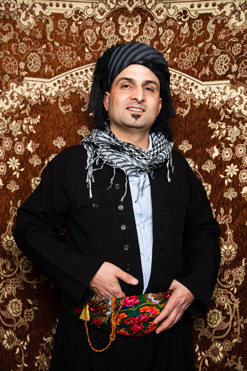 Portrait of refugee Rzgar wearing a turban and a kefiyah scarf around his neck holding onto a embroidered floral fanny pack