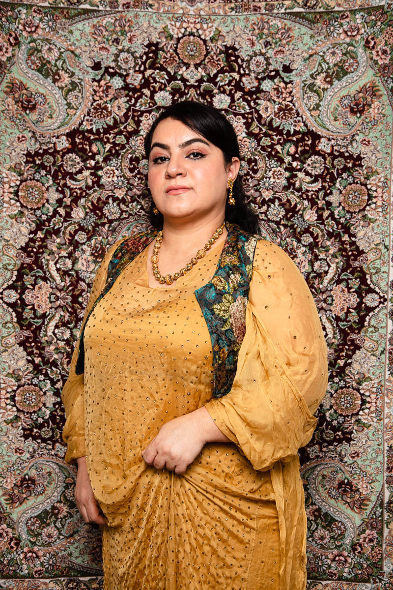 Portrait of refugee Shokhan wearing a golden sequined gown and floral waist coat standing against a traditional rug background