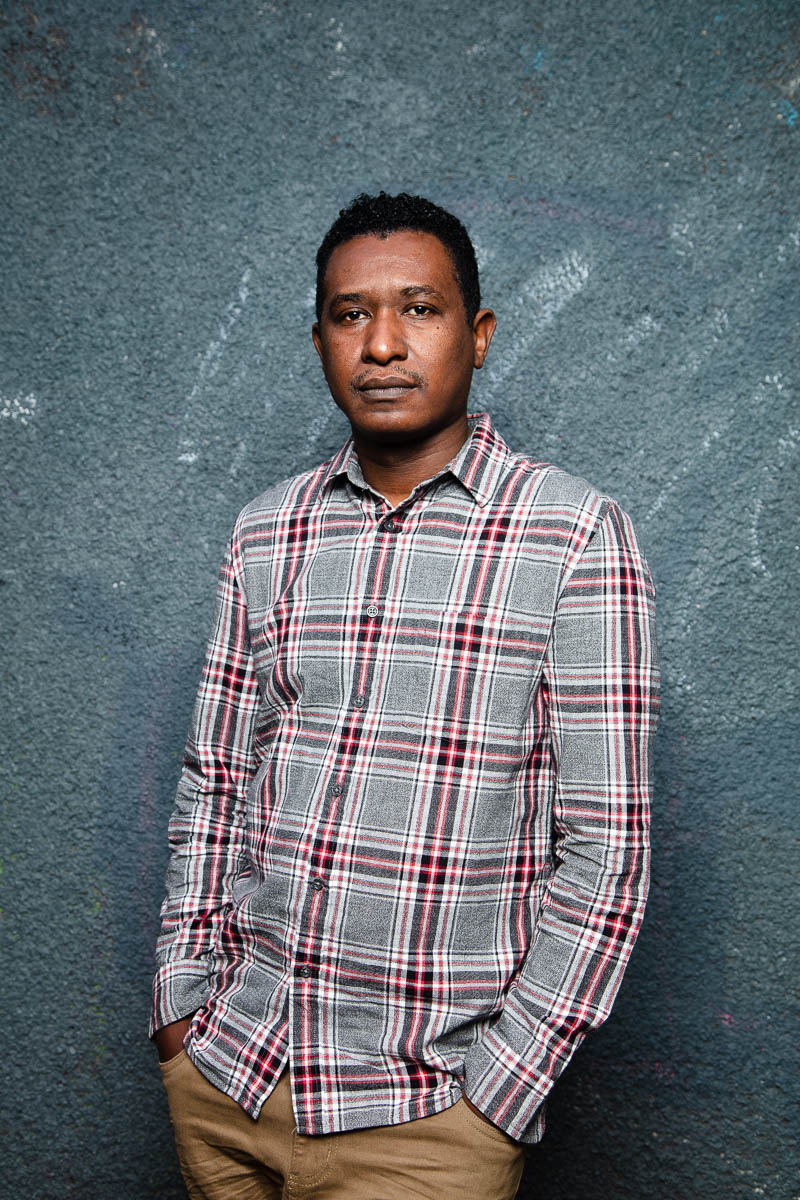 Portrait of refugee Ali wearing a plaid shirt with his hands in his pant pockets